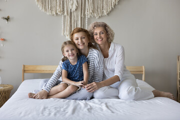 Family ties, love and heredity. Cute little girl her young mother and mature granny hugging seated on bed in cozy bedroom, staring at camera, enjoy priceless time together and their strong connection