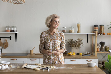 Pensive mature woman cook in kitchen, distracted from cookery process, staring into distance, smile, enjoy pleasant house chores, prepare dessert for family. Carefree retiree cooking cookies at home