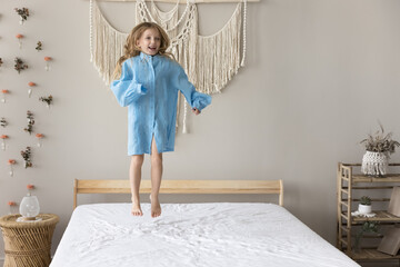 Little girl in blue parent shirt spend leisure at home in cozy bedroom, jump on fresh bed linen after waking up, have fun, smile, enjoy playtime, engaged in funny activity, relish carefree childhood
