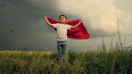 Happy child playing superhero against sky. Little hero in red cloak looks into distance. Boy kid...