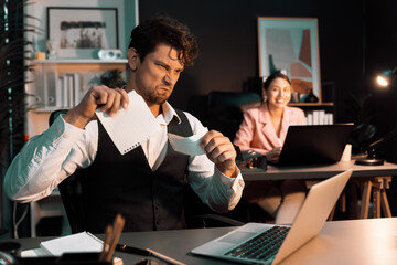 Sitting man with serious face tearing failure paperwork of business project at night lighting time at modern office with blurry secretary checking paper report with folder background. Postulate.