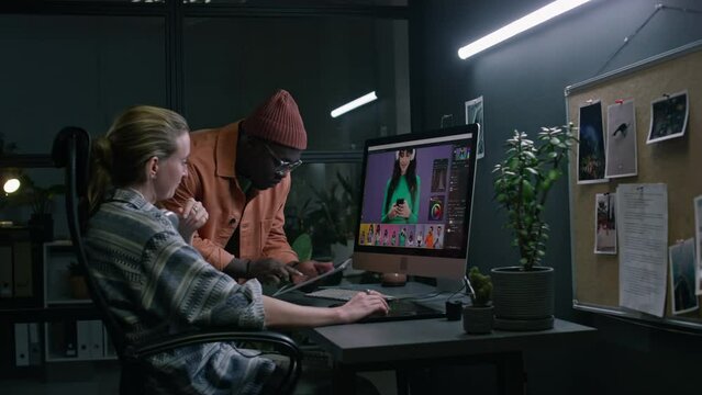 Medium side of Caucasian female graphic designer working on commercial images on computer in postproduction studio, African American male colleague showing reference on tablet, giving recommendations