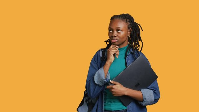 Pensive woman carrying laptop, doing thinking gesture, memorizing information for school exam, studio background. Student holding notebook, remembering before university assessment, camera A