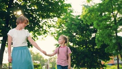 Child girl smiles walks through park with green trees, holding her mothers hand. Daughter child, mom holding hand, walking in park summer day. Family relations. Happy family resting together, nature