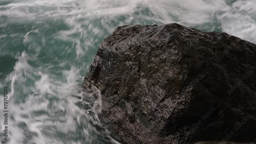 Poster A large rock is in the ocean with waves crashing around it. The water is a deep blue color and the rock is a dark grey color. Scene is calm and peaceful, as the rock stands strong against the waves
