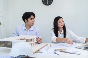 Professional male asian architect using ruler to measure house model length while young beautiful caucasian colleague using laptop to analyzed data on meeting table with house model. Immaculate.