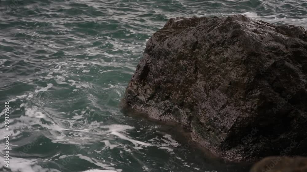Poster A large rock is in the ocean with waves crashing around it. The water is a deep blue color and the rock is a dark grey color. Scene is calm and peaceful, as the rock stands strong against the waves