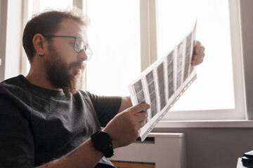 Focused bearded man with glasses looking at film negatives in bright natural light beside a window.