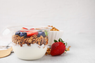 Tasty granola with berries and yogurt in plastic cup on light table, space for text