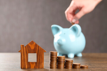 Woman putting coin into piggy bank at wooden table, focus on house model and money