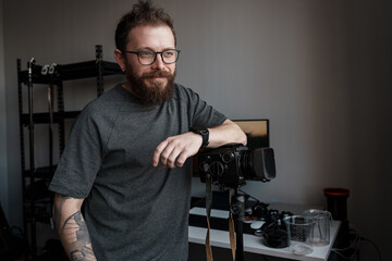Confident male photographer leaning on his camera on a tripod in a well-organized home studio setting.