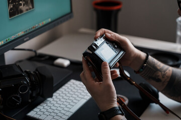 Close-up of a person's hands adjusting a digital camera, with a computer, keyboard, and another camera on a desk in the background.