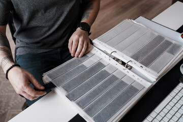 A person carefully examines film strips laid out in a binder on a office desk, epitomizing the meticulous nature of film photography. - obrazy, fototapety, plakaty