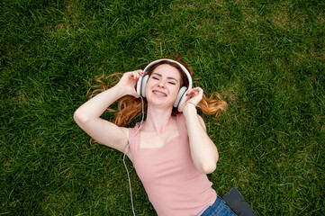 Top view of a young red-haired woman lying on the grass and listening to music on headphones. 