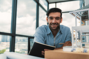 Smiling smart architect engineer looking at camera while sitting near house model. Happy confident businessman wearing casual outfit holding notebook while poses at camera. Civil engineering. Tracery.