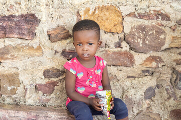 village african girl eating snacks, sited in the yard in front of the house