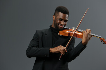 Talented young African American man in elegant black suit playing violin on grey background, music performance concept