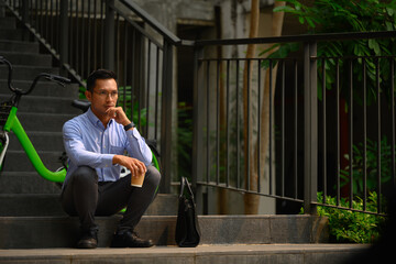 Thoughtful office worker guy looking away while sitting on stairs near office building - 797235948
