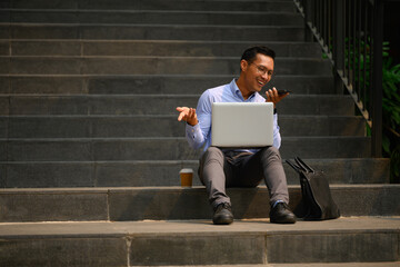 Full body shot of smiling young businessman talking on smart phone sitting with laptop on steps - 797236743