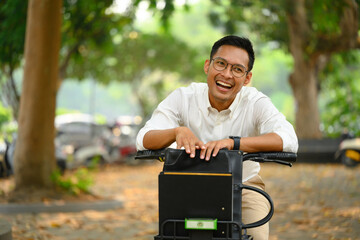 Cheerful Asian male office worker with bicycle in city park. Active lifestyle and transport concept