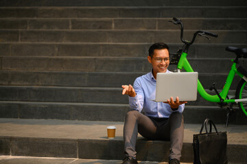 Cheerful male office worker sitting on the stairs in the city and using laptop