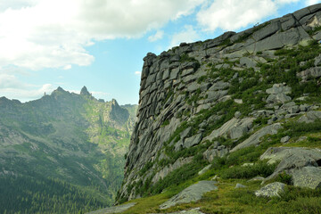 A high mountain with a sheer steep layered slope covered with grass overlooking rock formations on a sunny summer day.