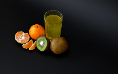 A glass of tropical fruit juice on a black background, next to pieces of ripe kiwi fruit and tangerines.