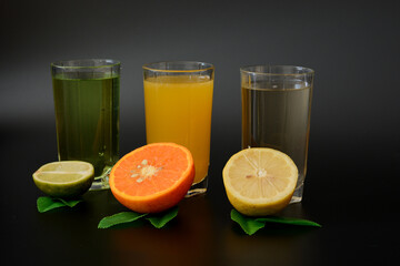 Three tall glasses with different citrus juices on a black background, next to the halves of ripe lemon, orange and lime with leaves.