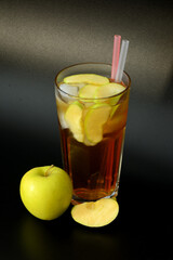 A fresh glass of fresh fruit juice with ice and straws on a black background, next to a ripe yellow apple.