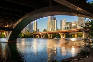 Beneath the North Lamar Bridge in Austin, Texas, the tranquil waters of Lady Bird Lake mirror the golden hues of the cityscape, creating a beautiful reflection on a summer day.