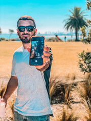 Handsome young Man Showing  Smartphone Screen. Young man in glasses standing on the beach with a City and palms as a background, Melbourne, Australia