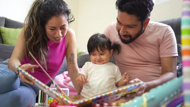 Happy hispanic family reading a learning book to their baby daughter, young latin couple enjoying time with their cheerful happy toddler