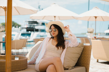 Beautiful young Asian girl is sitting in the cafe near the harbor while holding her straw hat with blurred yacht in the background. Wearing slim cream dress, pink and reflective sunglasses, necklace.