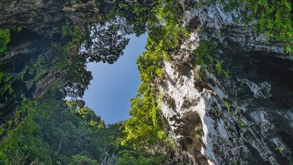 A bright blue sky surrounded by a frame of sheer cliffs. Green tropical vegetation grows on steep limestone slopes. A hole in the ceiling of the cave. Malaysia. Kuala Lumpur. Batu caves