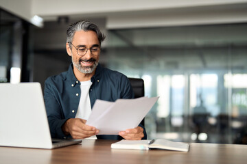 Happy mature professional business man checking documents working in office. Middle aged male executive, entrepreneur or manager sitting at desk reading corporate financial report contract. Copy space