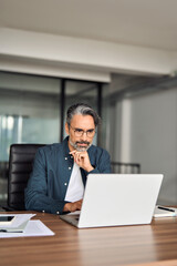 Busy older serious middle aged business man sitting at desk using computer. Mature businessman executive wearing shirt and glasses working looking at computer technology in office. Vertical photo.