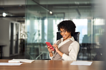 Happy busy young professional African American business woman executive holding cellphone using mobile looking at mobile cell phone tech with smartphone in hands sitting at desk in office working.