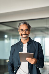 Vertical portrait of happy smiling older business man executive standing in office using digital tablet. Middle aged businessman manager wearing shirt glasses working on professional financial project