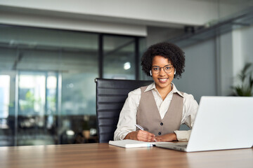 Happy young African American business woman wearing earbud working in office using laptop looking at camera advertising elearning, online learning webinars, virtual meetings with clients. Copy space.