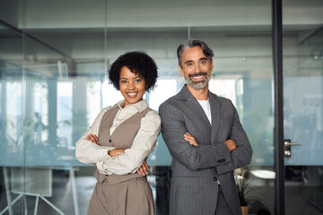 Two diverse business partners, employees or executives team standing in office arms crossed looking at camera. Business man and woman sales managers, company workers, professionals corporate portrait.