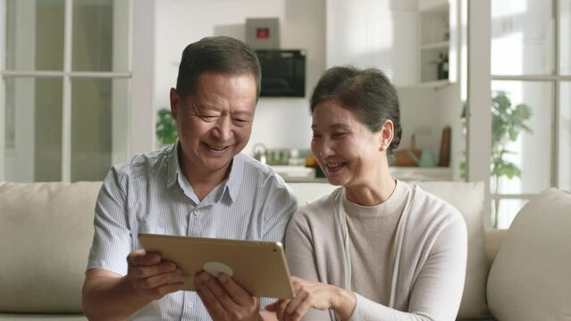 happy senior asian couple sitting on couch at home looking at digital tablet together