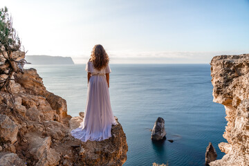 A woman in a white dress stands on a rocky cliff overlooking the ocean. The scene is serene and...