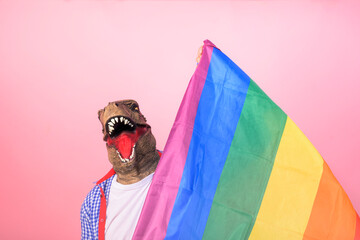 Man in T-Rex mask holding a rainbow flag looking at the camera on an isolated pink background