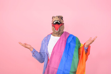 person in dinosaur mask with open arms holding a rainbow flag on an isolated pink background