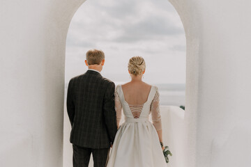A bride and groom are walking through a white archway on a sunny day. The bride is wearing a white dress and the groom is wearing a suit. The archway is decorated with flowers.