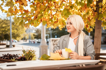 A blonde woman sits at a table with a cup of coffee and a leaf on it. The scene is set in a city with a tree in the background.