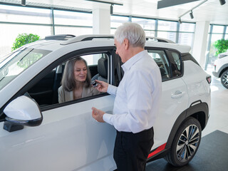 Mature Caucasian woman sits in a new car, an elderly man gives her the keys. 