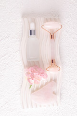 White matte bottle with natural remedy, serum, massage oil on white plaster embossed podium among flowers. Care concept. Vertical Top view. Blank lay