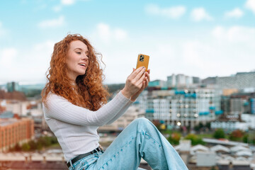 happy young lady with red curly hair exploring city and taking selfies on mobile phone
