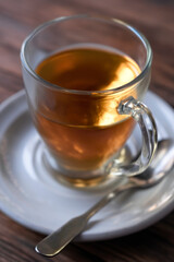 Glass filled with hot tea, on a white saucer with a spoon next to it on a wooden table. Narrow depth of field, focus on the handle